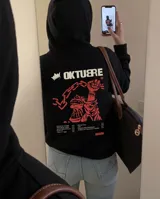Canguro negro con capucha y bolsillo frontal, confeccionado en tejido de algodón y poliéster con interior de felpa. Presenta un estampado gráfico en la espalda en color rojo y crema con motivos de cadenas y una mano alzada, junto con una lista de canciones y el logo de la marca.