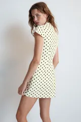 Vestido corto de color blanco con estampado de lunares negros. Posee cuello redondo con lazada, escote cuadrado en la parte delantera y abertura tipo gota con lazo en la espalda. Mangas cortas y silueta entallada.