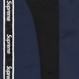 Pantalón deportivo Supreme de corte holgado, color azul marino, con franjas laterales negras que incluyen el logo de la marca repetido. Presenta cintura elástica y puños ajustados en los tobillos.