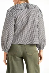 Blusa de manga larga con estampado de cuadros vichy en blanco y negro. Presenta un cuello con volados amplios y un detalle de lazo negro en el centro del escote.