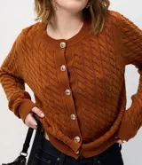 Cárdigan de punto trenzado color marrón, con cuello redondo, manga larga y cierre frontal con botones metálicos. Presenta puños y ruedo acanalados.