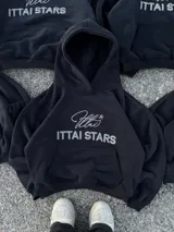 Hoodie de color negro con diseño de estampa frontal que combina una tipografía manuscrita con el texto ITTAI STARS en letras mayúsculas. Presenta un corte holgado, capucha amplia y bolsillo tipo canguro.