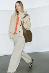 Trench corto color verde militar, con cuello solapa y capucha. Tiene manga larga con trabillas y botón en los puños, bolsillos delanteros y bajo asimétrico. Cierre frontal con botones. El tejido es repelente al agua.