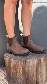 Bota tipo Chelsea de cuero marrón con textura granulada, paneles laterales elásticos en color negro y suela track dentada con costura blanca visible.