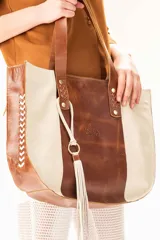 Bolso shopper de cuero vacuno en combinación de tonos arena y whisky. Presenta un diseño amplio con detalles de trenzado artesanal en los laterales, asas largas para llevar al hombro y un colgante decorativo con flecos de cuero. Incluye logo grabado de la marca Lorena Caprile.