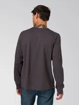 Remera de manga larga color gris oscuro, con cuello redondo y puños acanalados. Presenta un pequeño parche rectangular con el logo de la marca en el lado izquierdo del pecho.