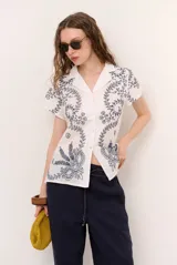 Camisa blanca de manga corta con cuello solapa, cierre frontal de botones y diseño bordado en hilo azul marino con motivos florales y vegetales.