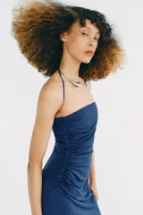 Vestido midi de color azul, con escote recto y cuello halter que incorpora una aplicación metálica tipo choker. Presenta un diseño con frunces laterales en el frente.