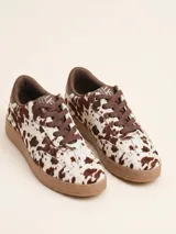 Championes urbanos con diseño de estampado animal print tipo vaca, confeccionados en cuero con textura de pelo. Presentan cordones marrones, suela de goma en tono tostado y detalles en color marrón oscuro en el cuello y lengüeta.