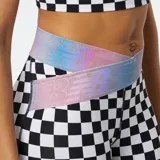 Calza deportiva New Balance con estampado a cuadros blancos y negros, cintura alta cruzada con diseño iridiscente y logo de la marca en la pierna.