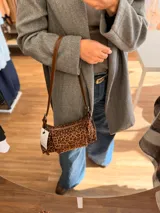 Cartera tipo baguette con estampado animal print, confeccionada en material suave símil gamuza. Presenta un bolsillo frontal con solapa y cierre, compartimento principal con cierre superior, y asa larga regulable de cuero sintético marrón.