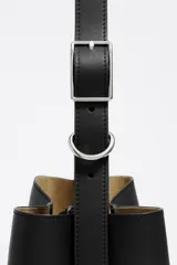 Bolso de hombro tipo cubo de estructura rígida, color marrón oscuro, confeccionado en piel vacuna. Posee asa regulable con hebilla metálica y cierre de cordón en la parte superior. Lleva el logo metálico de Bimba y Lola en la parte frontal inferior.