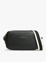 Cartera tipo bandolera Tommy Hilfiger de color negro, con acabado liso, diseño compacto y esquinas redondeadas. Cuenta con cierre de cremallera superior, inscripción y logotipo de la marca en bajo relieve dorado en el frente, y una correa cruzada ancha ajustable de jacquard a rayas en blanco y negro con la marca escrita.