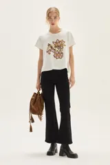 Remera blanca de algodón con cuello redondo y manga corta. Presenta un estampado bordado central con motivos florales y un animal (posiblemente un conejo o liebre) en tonos lila, morado y marrón.