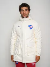 Parka larga de abrigo color crema con capucha, bolsillos frontales con solapa y cierre frontal. Presenta el logo de Umbro y el escudo del Club Nacional de Football bordados en el pecho.
