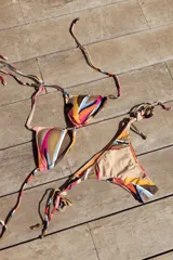 Bikini de dos piezas con estampado abstracto en tonos tierra, naranja, rosa y azul. El top es de triángulos movibles con almohadillas desmontables y se ata al cuello y espalda. La bombacha es vedetina con tiritas ajustables a los costados.