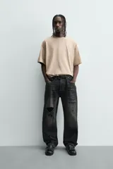 Camiseta de manga corta y cuello redondo, color beige claro, con corte relajado (relaxed fit) y un pequeño texto bordado en el pecho.