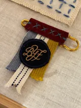 Pin decorativo con estructura de gancho metálico dorado, compuesto por una tira de cuero bordó con costuras en cruz y tres tiras de tela en tonos azul, crema y mostaza. Incluye un parche circular negro con las iniciales BYF bordadas en hilo color ocre.