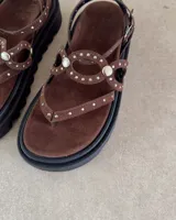 Sandalia de plataforma negra con tiras decoradas con tachas metálicas plateadas. Posee un diseño tipo ojota con ajuste de pulsera al tobillo mediante hebilla.
