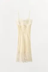 Vestido midi de estilo lencero confeccionado en viscosa satinada color crema. Presenta escote recto con tirantes finos, detalle de encaje floral en el pecho y el ruedo, y pliegues verticales en el frente.