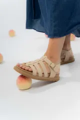 Sandalia estilo gladiadora de color beige, confeccionada en material gamuzado sintético. Presenta múltiples tiras anchas que sujetan el pie, ajuste lateral con hebilla metálica y suela plana antideslizante de color marrón.