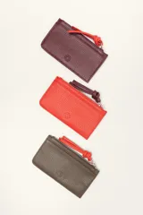 Monedero rectangular de cuero vacuno con textura granulada en color marrón topo. Cuenta con cierre superior metálico y un tirador decorativo de cuero en color naranja con un nudo. Presenta un pequeño logo grabado en el centro.