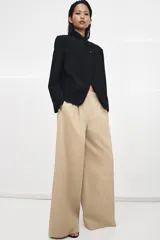 Pantalón de tiro medio con corte wide leg, confeccionado en algodón. Presenta cintura con trabillas, bolsillos laterales ocultos y detalle de pliegues en el delantero. Cierre frontal con cremallera, botón y ganchos metálicos.