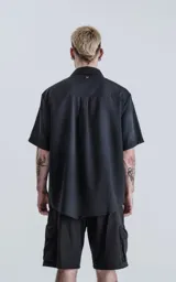 Camisa de manga corta en color negro con proceso de lavado, cuello clásico y cierre frontal con botones. Presenta un diseño minimalista con una costura vertical decorativa en el frente.