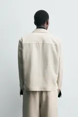 Sobrecamisa de hombre color beige claro, con cuello de solapas de pico, manga larga y cierre frontal de botonadura. Presenta bolsillos de vivo en la cadera.