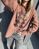 Camisa de algodón con corte clásico, cuello camisero y cierre frontal con botones. Presenta un diseño bordado en punto de cruz con motivos florales en color bordó sobre el frente. Cuenta con mangas largas con puños abotonados.