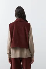 Chaleco de pana color marrón oscuro, con cuello camisero, cierre frontal de botones metálicos dorados y dos bolsillos frontales con detalle de bordado geométrico.