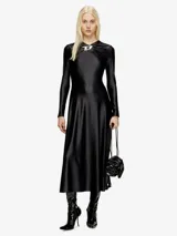 Vestido largo negro de manga larga y cuello drapeado, con broche metálico en forma de 'D' en el escote. El vestido tiene un corte al cuerpo en la parte superior y cae en una falda fluida.