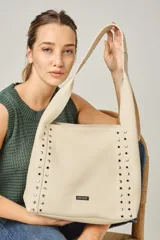 Cartera tipo tote de cuero vacuno color natural, con diseño de ojalillos metálicos en los laterales frontales y logo de la marca en placa metálica. Cuenta con manija única para llevar al hombro.