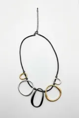 Collar Galaxia de diseño asimétrico, con cordón negro y dijes colgantes en forma de argollas y óvalos de acabado mate en tonos plateado, dorado y negro. Incluye cadena de extensión metálica.