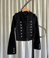 Campera estilo militar de color negro, con cuello mao y cierre frontal. Presenta múltiples botones metálicos plateados dispuestos en filas horizontales y detalles de charreteras en los hombros. Las mangas cuentan con puños decorados con botones a juego.