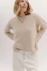 Buzo de lambswool color beige, con escote redondo y detalle en V. Presenta cierres metálicos en los puños que revelan un interior en contraste de color marrón y ribete rojo. Tiene tajos laterales para mayor comodidad y un fit amplio.