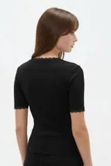 Remera de algodón negro con textura acanalada, manga corta y cuello redondo base. Presenta sutiles detalles de puntilla en el cuello y en los bordes de las mangas.