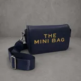 Cartera tipo bandolera de tamaño pequeño, color azul marino con textura granulada. Presenta el texto 'THE MINI BAG' estampado en letras doradas en el frente y cuenta con una correa de cinta ajustable al tono con herrajes metálicos plateados.