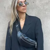 Riñonera negra de cuero sintético con tres bolsillos frontales con cierre plateado y correa ajustable con hebilla.