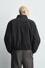 Cazadora de hombre de ajuste cropped fit, confeccionada en tejido de algodón con patrón de rayas verticales finas en tonos oscuros (negro/azul marino y marrón/beige). Presenta cuello solapa, cierre frontal de cremallera, bolsillos laterales y elástico en puños y bajo.