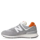 Championes urbanos New Balance modelo 574, color gris con detalles en blanco y naranja en el talón. Confeccionados en gamuza y malla, cuentan con el logo N característico en los laterales y entresuela con tecnología ENCAP para mayor amortiguación.