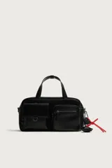 Bolso bandolera negro de acabado liso, con compartimento principal con cierre de cremallera. Cuenta con dos bolsillos frontales, uno con solapa y detalle metálico y otro con cremallera. Incluye dos asas de mano y un asa de hombro ajustable y extraíble, además de detalles de charms y mosquetón en un lateral.