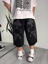 Bermuda baggy negra de denim con bolsillos traseros amplios y bordados contrastantes en hilo blanco con diseño de cruces y motivos barrocos.
