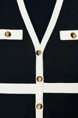 Cárdigan de punto en color negro con detalles en blanco en los bordes, escote en V y cierre frontal con botones metálicos dorados. Presenta mangas largas y bolsillos decorativos en el pecho.
