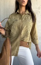 Camisa oversize de color verde oliva, con cuello clásico y cierre frontal de botones. Presenta un diseño bordado detallado en la parte superior delantera y mangas largas con puños.