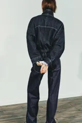 Cazadora de denim azul oscuro con cuello solapa, cierre frontal con cremallera y botones a presión ocultos. Presenta mangas largas tipo murciélago con puños, bolsillos de vivo en el delantero y bajo elástico ajustable con cordones.