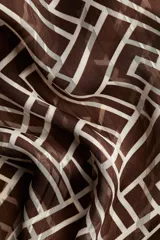 Blusa de manga larga en tejido transparente con estampado geométrico en tonos marrón y blanco. Presenta cuello elevado con frunces y puños ajustados con abertura y botón.