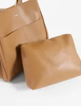 Bolso tipo shopper color marrón claro, de cuero sintético, con doble asa larga para llevar al hombro y bolsillo externo. Incluye estuche interno extraíble.