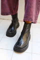 Bota de caña media color negro con textura de reptil en el empeine y paneles laterales elásticos. Presenta una suela de plataforma gruesa y punta cuadrada.