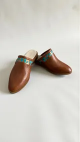 Sandalias tipo mules color marrón terracota, confeccionadas en cuero, con punta redonda, suela plana y detalles pintados a mano en tonos turquesa y dorado con aplique metálico triangular en el lateral.